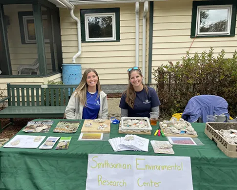 Two women sitting behind a table that is covered with pamphlets, magazines, and oyster shells. A sign on the table says Smithsonian Environmental Research Center. Behind the table is a house.